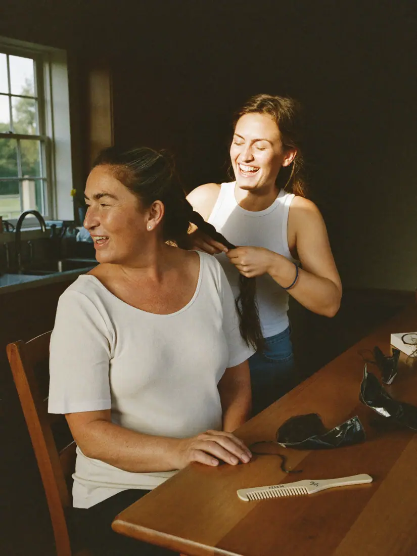 Two friends in a kitchen, one braiding the other's hair.